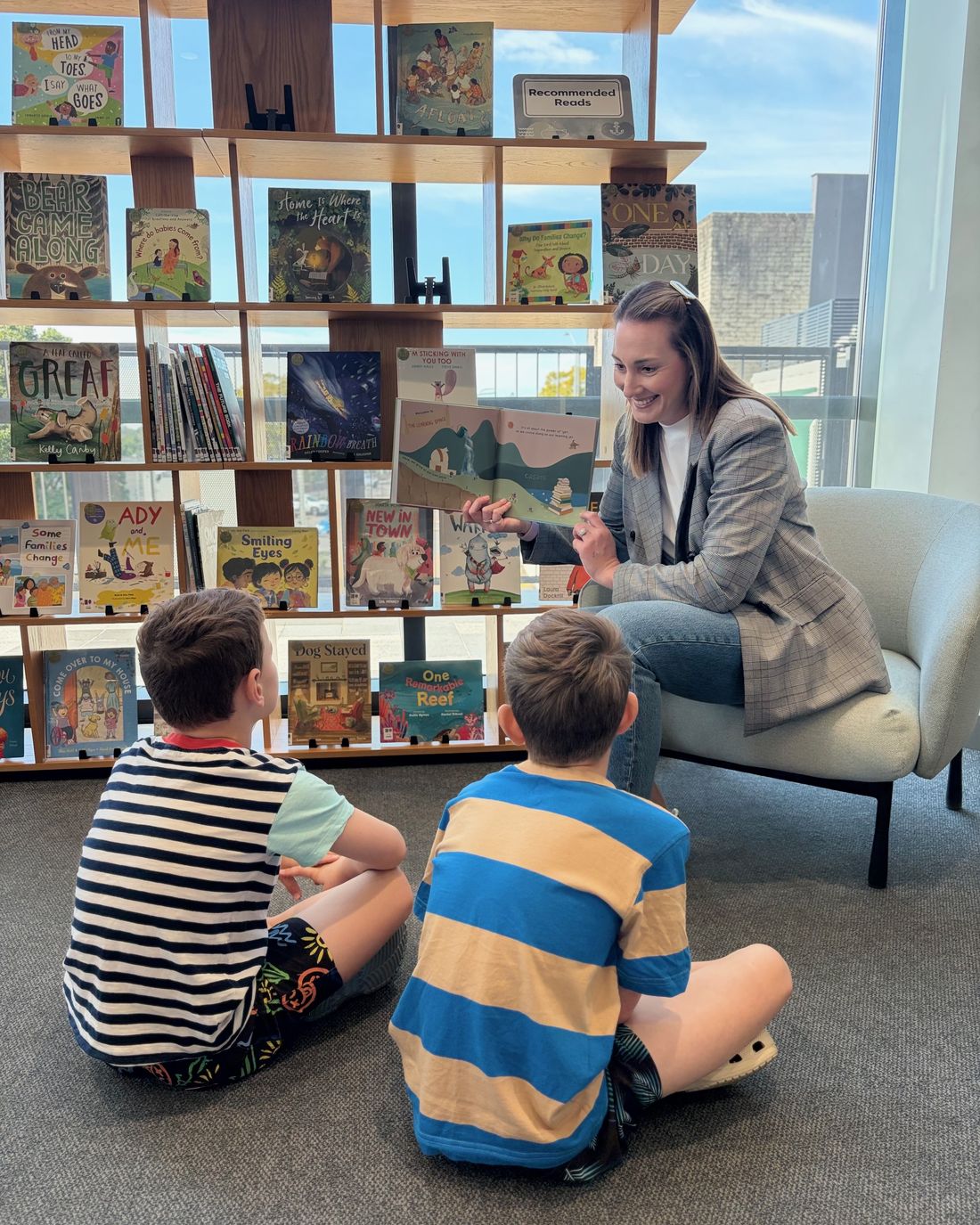 Hannah reading The Yet Jet aloud to two children in a library, holding the open book up for them to see.